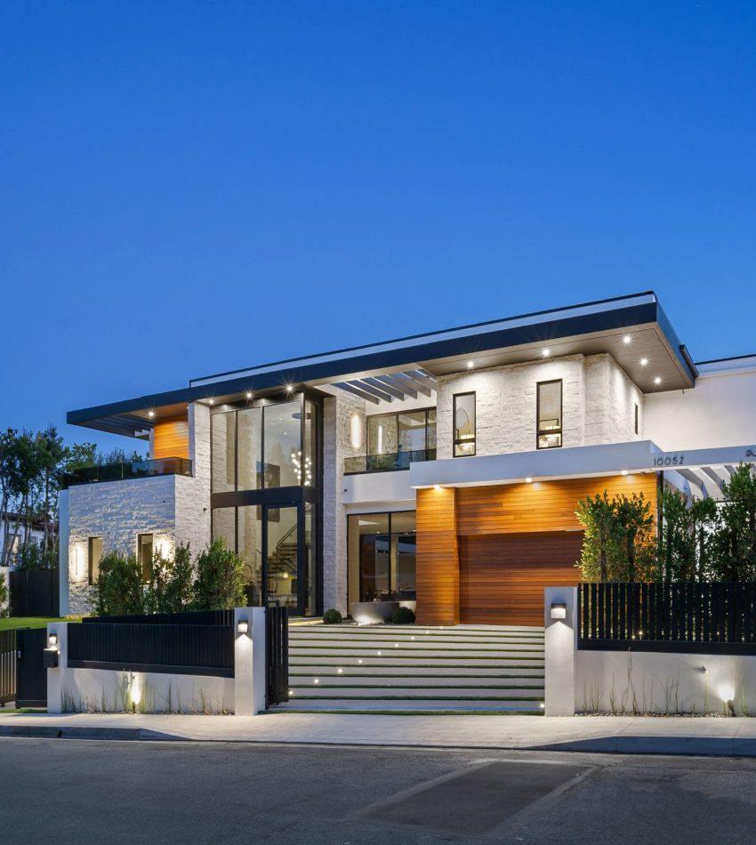 Modern luxury house exterior with large windows, illuminated at dusk, surrounded by greenery.